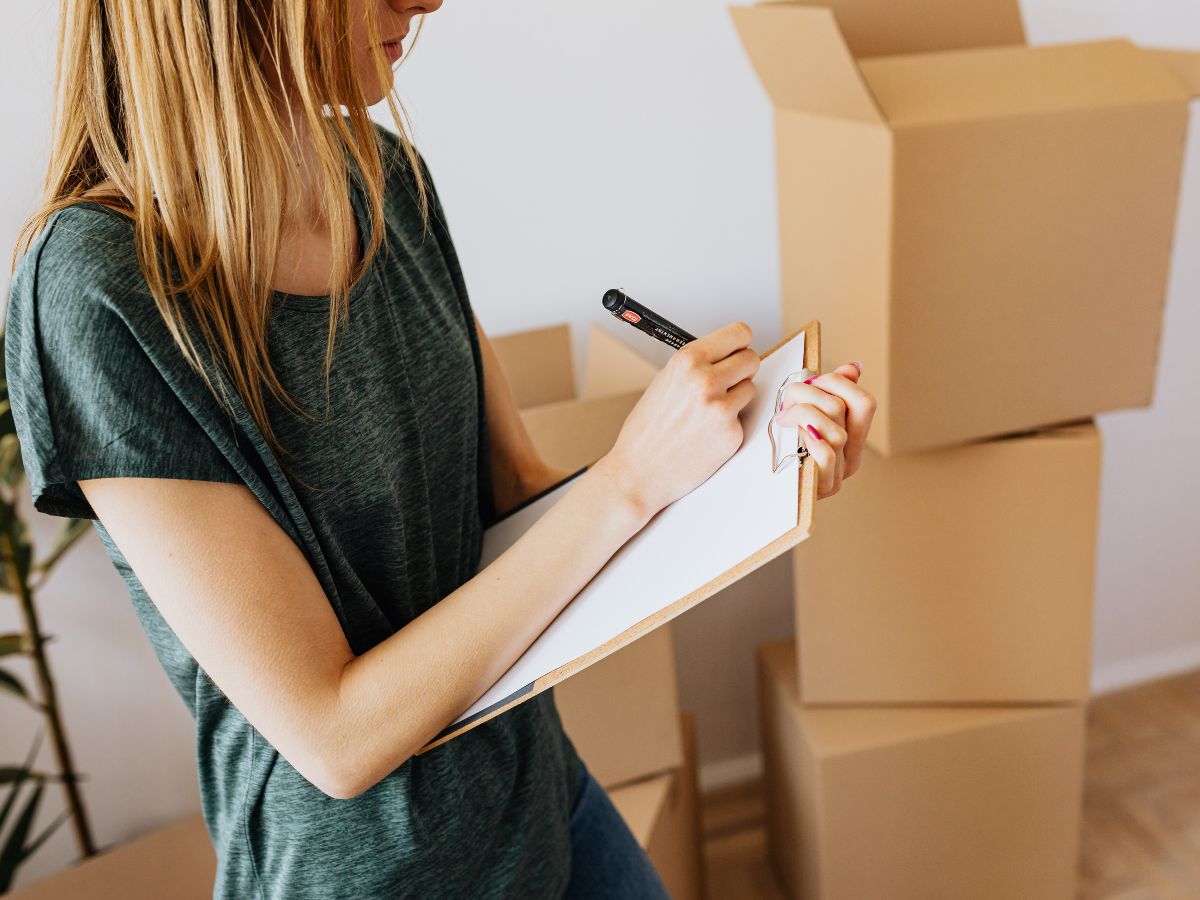 Szőke, hosszú hajú nő zöld felsőben listát ír, háttérben papírdobozok