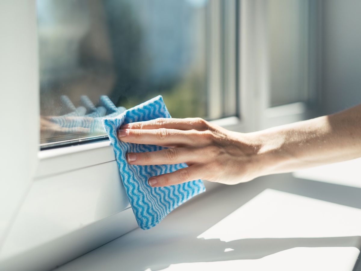 Kéz közelről, kék törlőkendővel tisztít egy fehér műanyag ablakkeretet és üveget, napfényben, finom mozdulatokkal eltávolítva a szennyeződést.