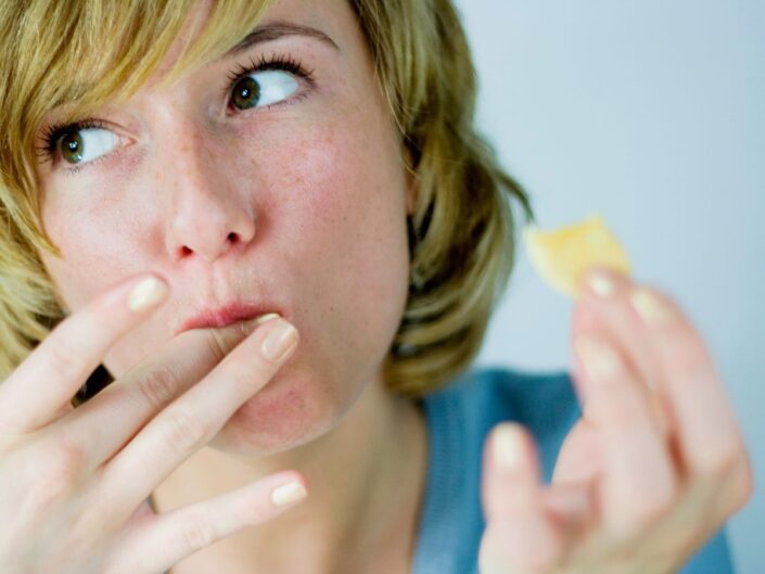 Közeli képen fiatal nő süteményt tart a kezében, másik kezének ujját lenyalja