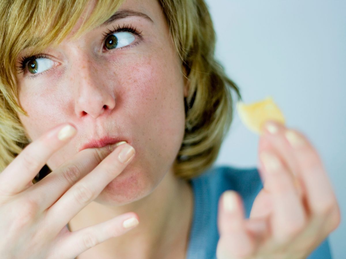 Közeli képen fiatal nő süteményt tart a kezében, másik kezének ujját lenyalja