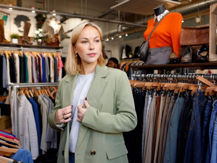 SZőke, fiatal, szép nő zöld blézert próbál fel használtruha-boltban
