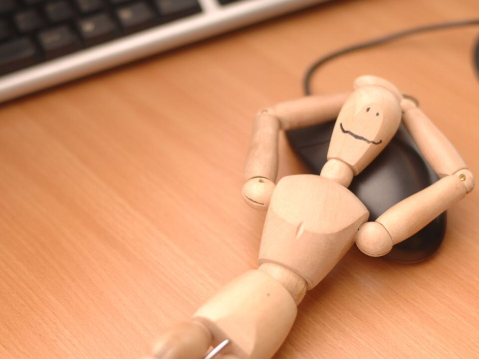 allando-faradtsag-nok-35-felett - Nők Lapja Egy fa bábu hanyatt fekszik egy számítógépes egér mellett az asztalon, kezeit a feje mögé teszi. A kép a kimerültség és passzív pihenés hangulatát idézi.