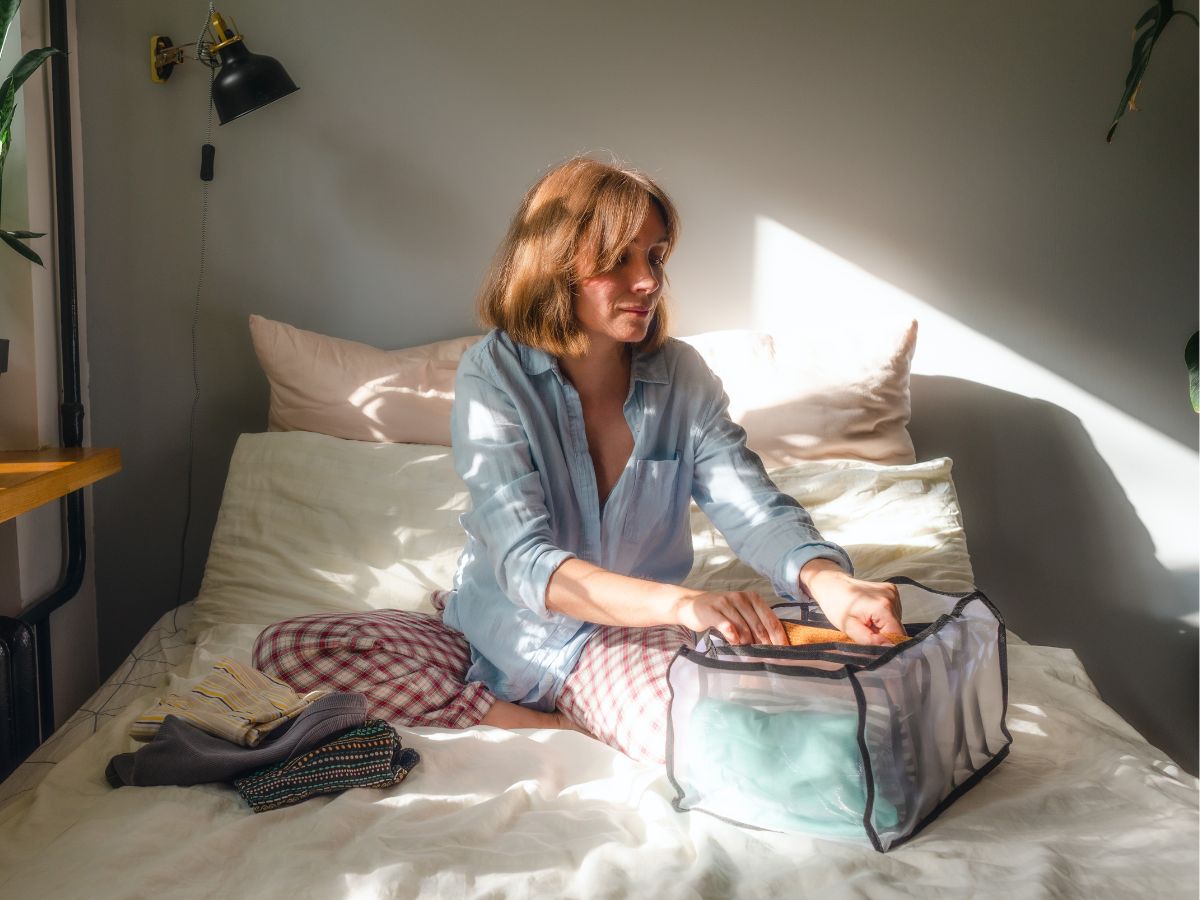 Nő ágyon ülve ruhákat pakol lezárható tárolókba a reggeli napfényben