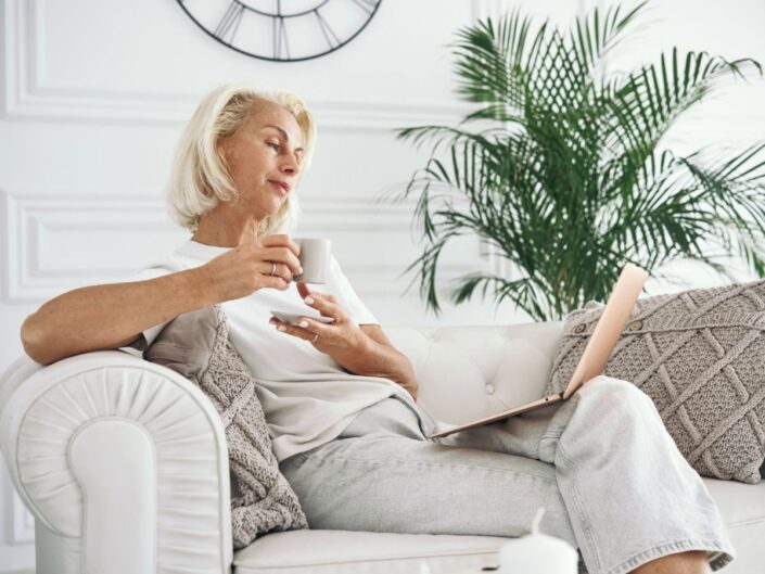 idősebb nő ül a kanapén kávéval és laptoppal