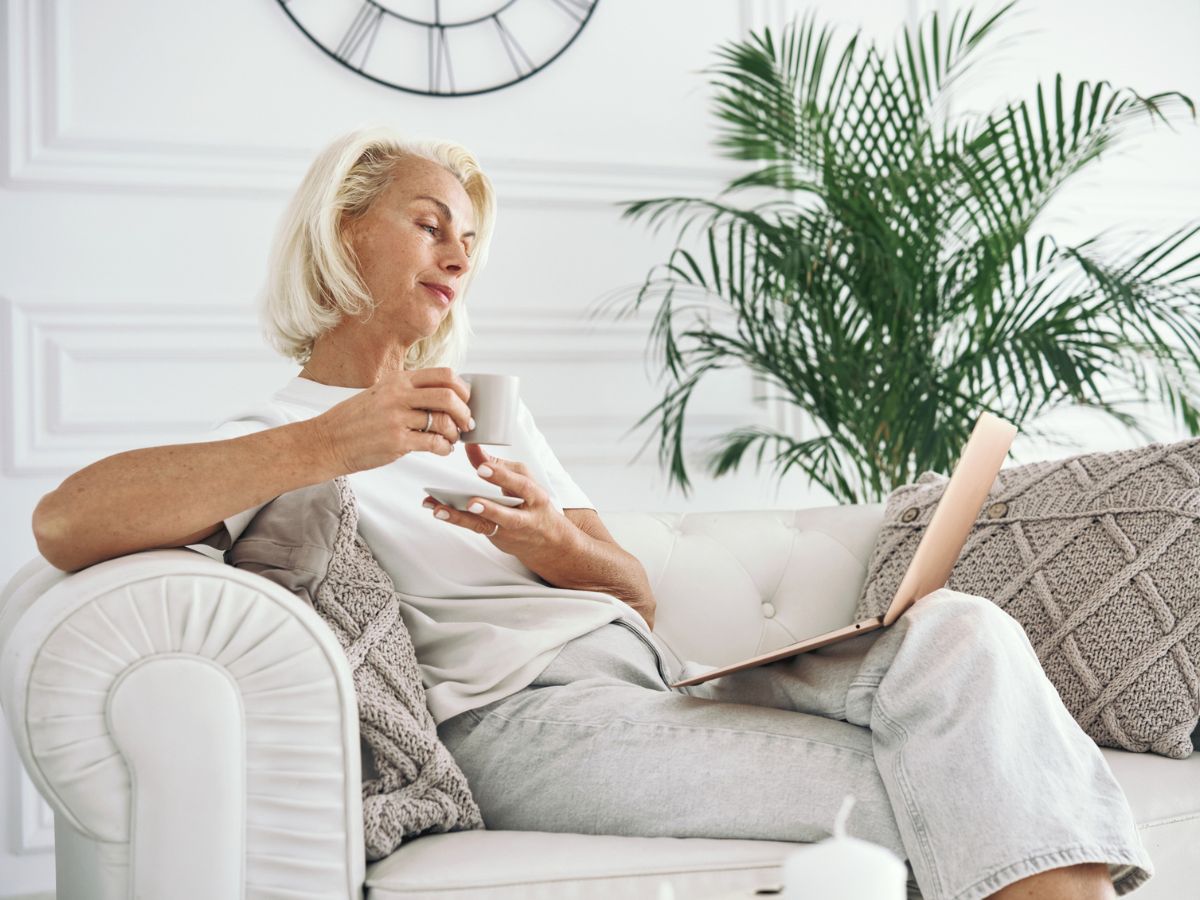 idősebb nő ül a kanapén kávéval és laptoppal