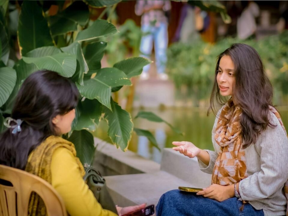 két hosszú hajú sálat viselő nő beszélget a parkban a víz mellett