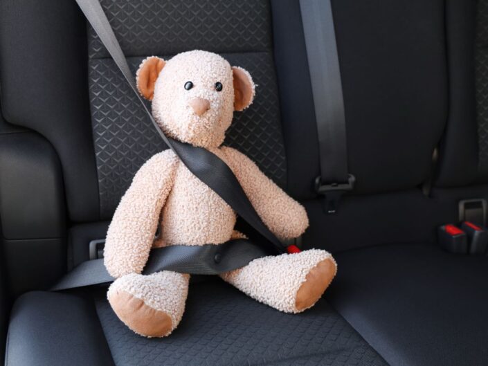 Egy plüssmackó ül az autó hátsó ülésén, biztonsági övvel bekötve; az öv átlósan fut a testén, a sötét kárpit kontrasztot ad a világos, puha játéknak.