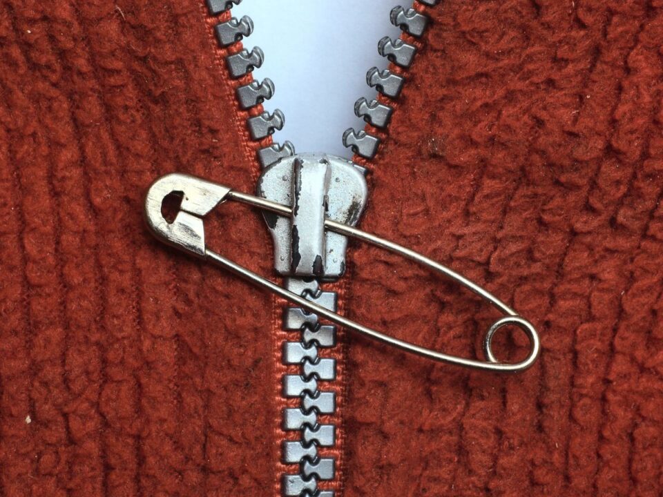 Barna kötött anyagon futó cipzár, amelyet egy biztosítótű tart össze ideiglenesen; a csúszka kopottnak tűnik, a fogak nem zárnak megfelelően.