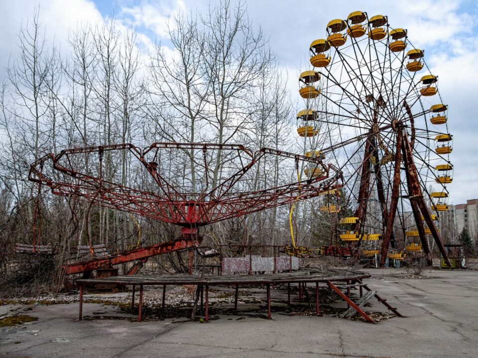 Rozsdás vidámparki játékok egy elhagyott térben: előtérben körhinta, háttérben sárga kabinos óriáskerék. A környéket benőtte a növényzet, a hely teljesen kihalt.