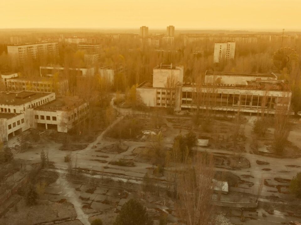 Madártávlati kép egy elhagyott városról: szürke lakóépületek, üres utcák és elvadult növényzet látható, a háttérben halványan kirajzolódik egy óriáskerék.