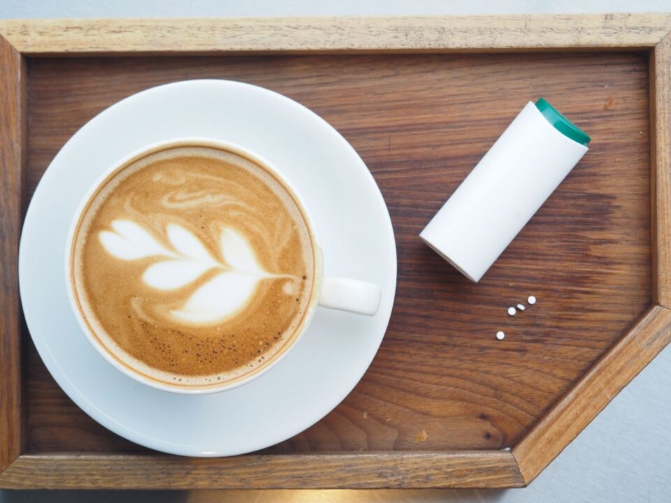 Egy cappuccino látható csészében, mellette kis fehér édesítő tabletták és egy adagoló egy fa tálcán. Letisztult, otthonos hangulatú jelenet felülnézetből.