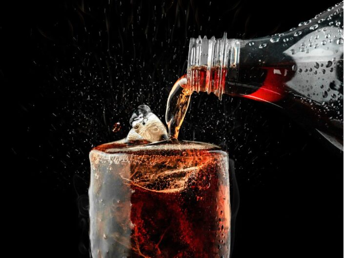 Egy pohár jéggel teli barna szénsavas üdítőbe éppen öntenek egy palackból. A buborékok pezsegnek, a folyadék fröccsen, sötét háttér előtt látványos, hűsítő hatást kelt.