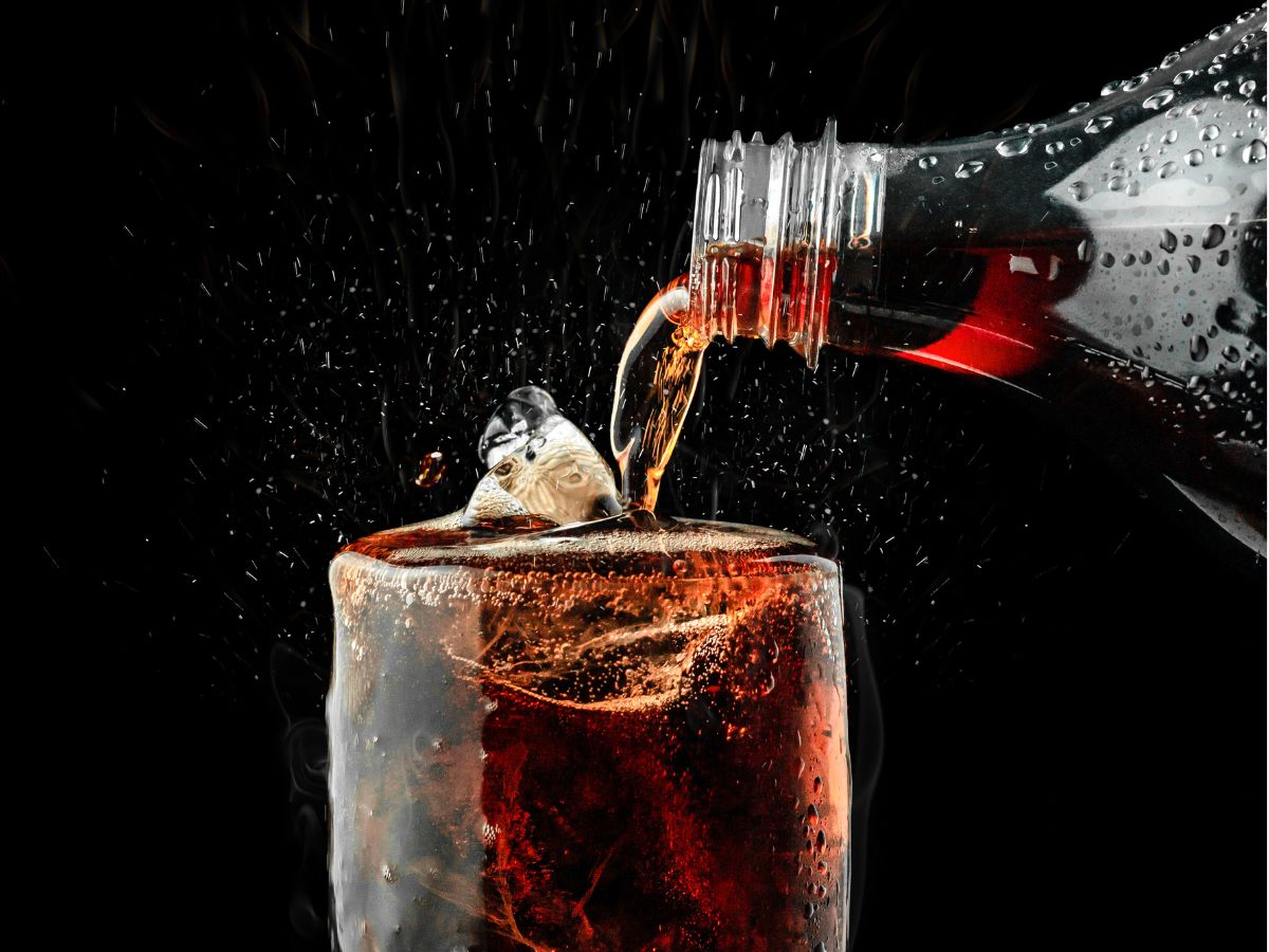 Egy pohár jéggel teli barna szénsavas üdítőbe éppen öntenek egy palackból. A buborékok pezsegnek, a folyadék fröccsen, sötét háttér előtt látványos, hűsítő hatást kelt.