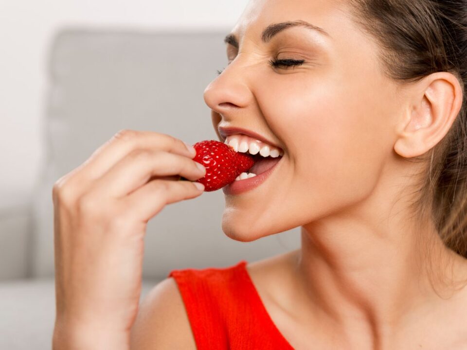 Egy nő közelről látható, amint mosolygva egy friss, piros epret tart a szájához, éppen beleharapni készül, természetes, világos háttér előtt.