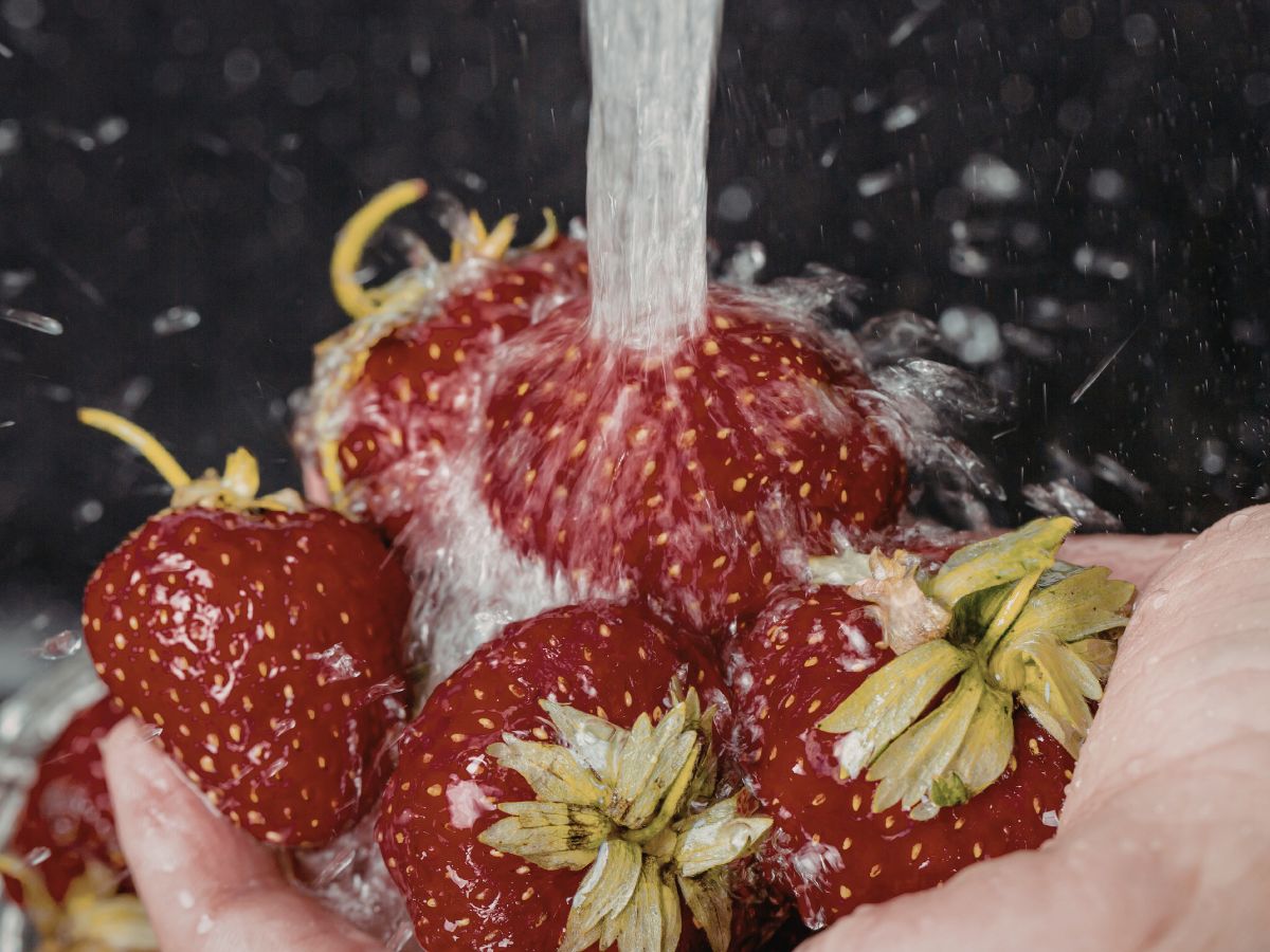 Friss epreket tartanak kézben, miközben erős vízsugár alatt mossák őket; a vízcseppek szétfröccsennek a gyümölcsökön, a zöld levelek még rajtuk vannak.