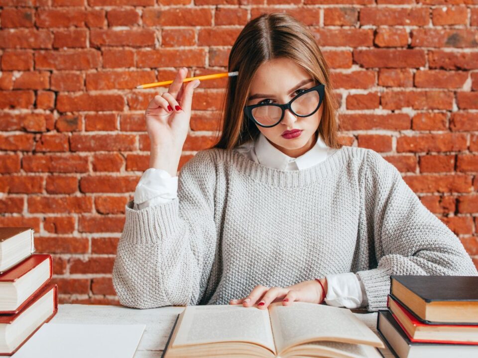 Egy fiatal nő szemüvegben könyv fölé hajol, ceruzát tart a kezében. Elmélyülten gondolkodik, körülötte könyvek, mögötte téglafal látható.