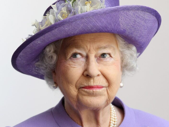 II. Erzsébet királynő közeli képen lila, virágos kalapban és ugyanilyen színű blézerben mosolyogva néz felfelé