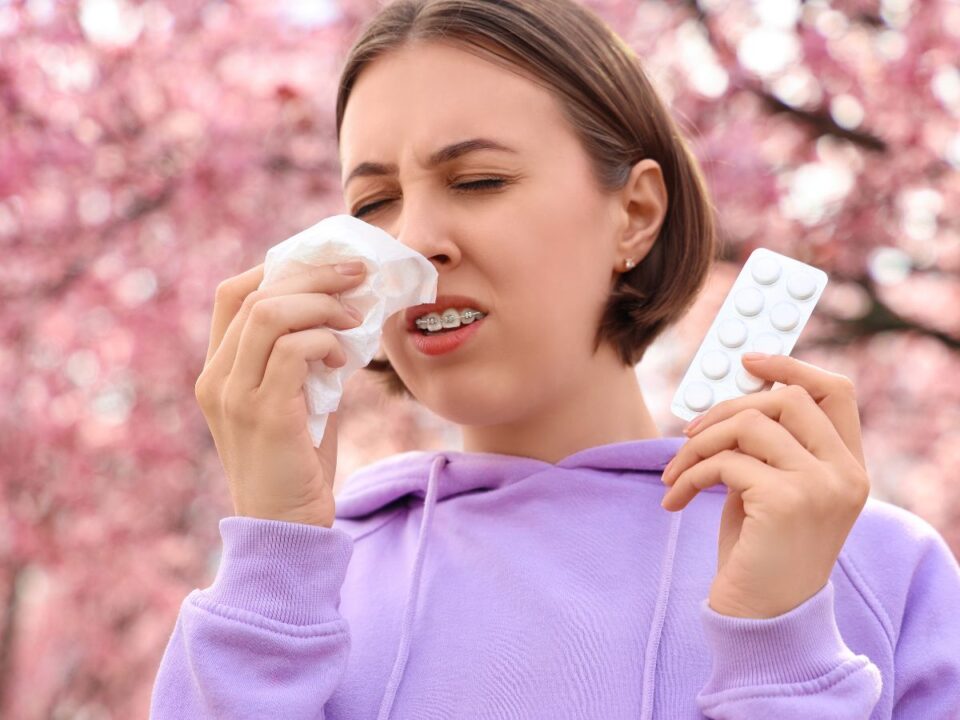 Fiatal nő virágzó fák előtt zsebkendőbe tüsszent, kezében gyógyszeres levél. Arca feszül, szeme csukva, a tavaszi pollen okozta allergiás tünetek jól láthatók.