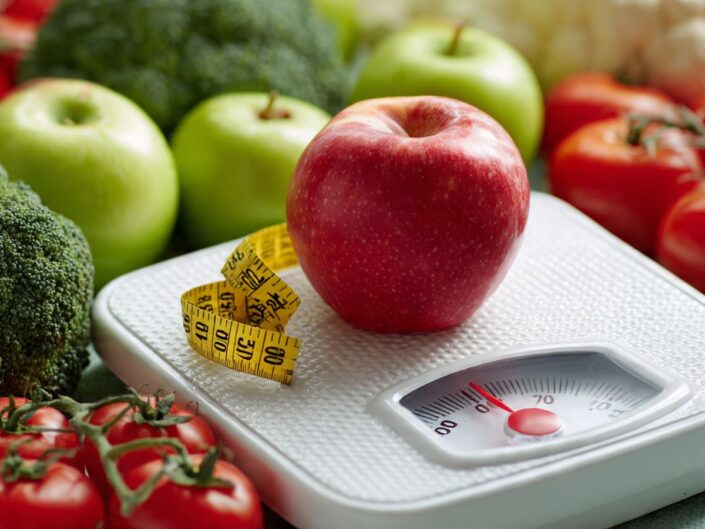 Egy mérlegen piros alma látható, mellette sárga mérőszalag, körülötte zöldségek és gyümölcsök. A kép az egészséges étkezés és a testsúly kapcsolatára utal.