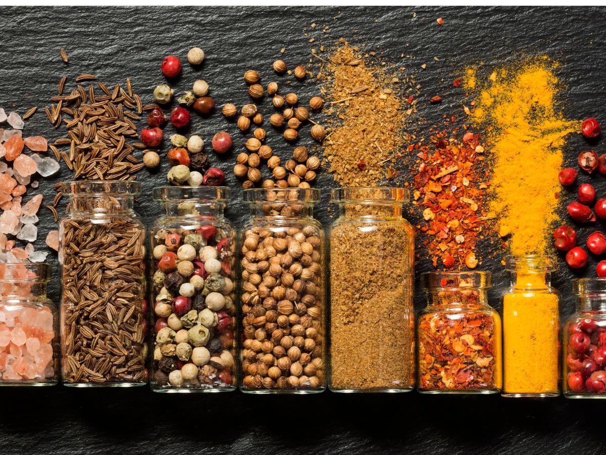 Különböző fűszerek üvegekben és kiszórva egy sötét felületen: kömény, bors, koriander, chili és kurkuma élénk színekben, egymás mellett rendezve.