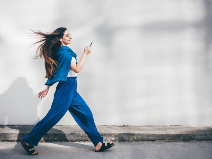 nő kék ruhában mobiltelefonnal sétál