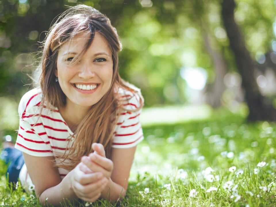 Egy mosolygó nő fekszik a fűben napsütésben, körülötte apró virágok; nyugodt, felszabadult hangulatot sugároz, mintha gondtalanul élvezné a pillanatot.