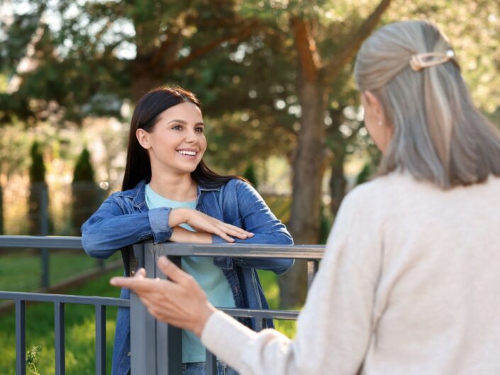 Két nő beszélget egy kertben kerítés mellett; a fiatalabb mosolyogva hallgatja az idősebb nőt, aki gesztikulálva mesél neki, barátságos, nyugodt hangulatban.