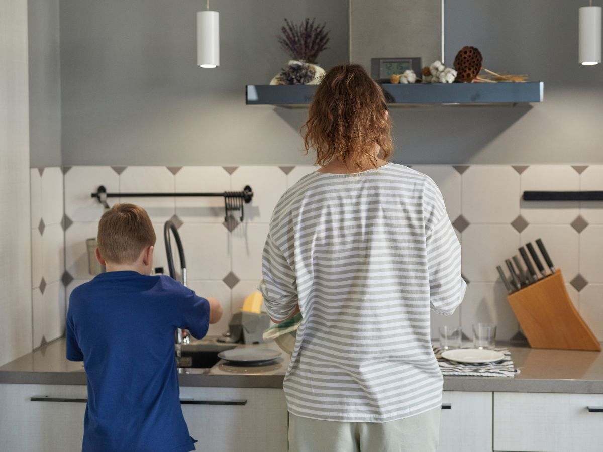 Egy kisfiú és egy felnőtt a konyhában mosogatnak, háttal állnak, közösen végzik a napi feladatokat rendezett, modern környezetben.