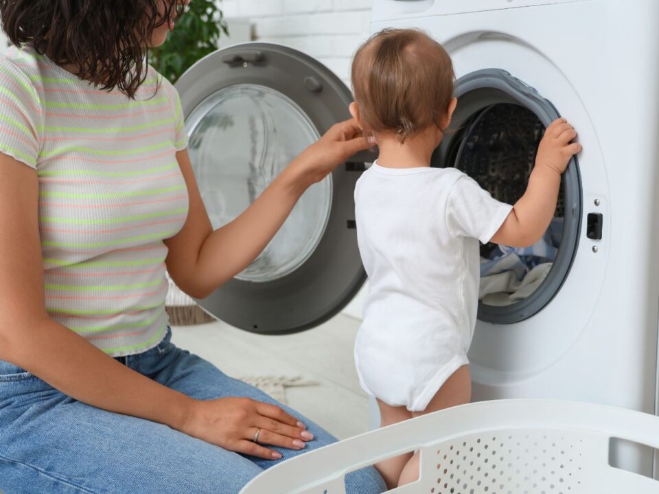 Egy kisgyerek egy elöltöltős mosógépbe pakol ruhákat, mellette egy felnőtt segít neki, így együtt végzik a házimunkát világos fürdőszobában.