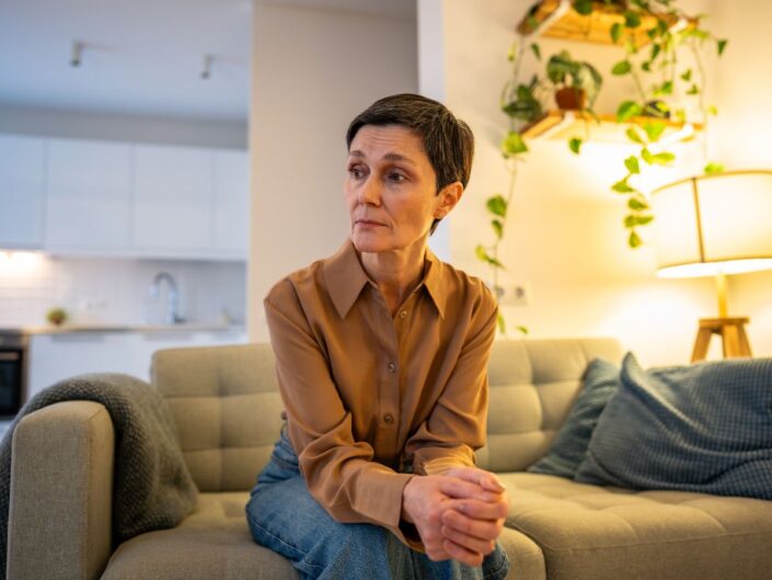 Egy rövid hajú nő a kanapén ül, kezét összekulcsolja, kissé oldalra néz. Arca feszült, a háttérben meleg fények és szobanövények látszanak, a hangulat csendes, elmélkedő.