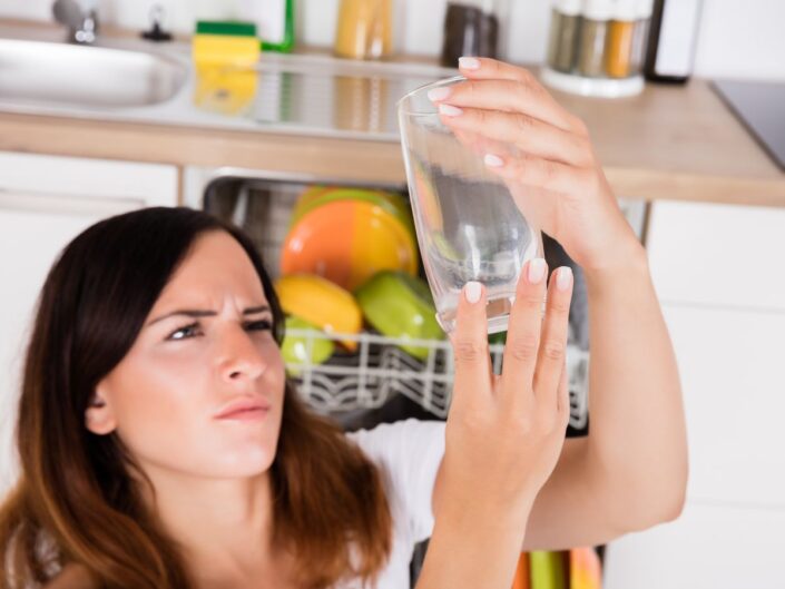 Egy nő a konyhában a mosogatógép előtt áll, és egy zavaros, foltos üvegpoharat tart a fény felé, miközben az alsó kosárban színes edények láthatók.