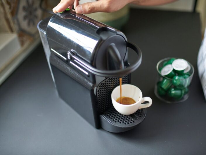 Fekete kávéfőzőgép szállodai szobában, fehér pohárba főzi a kávét