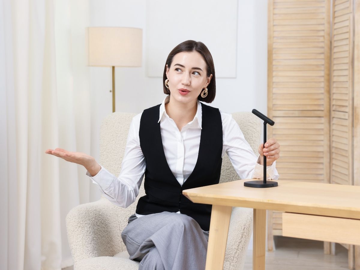 fiatal nő előadást tart asztalnál ülve mikrofonba beszélve fehér blúzban