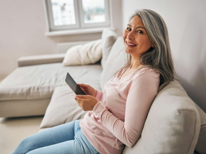Hosszú, ősz hajú intelligens nő kényelmes ruhában ül otthona kanapéján