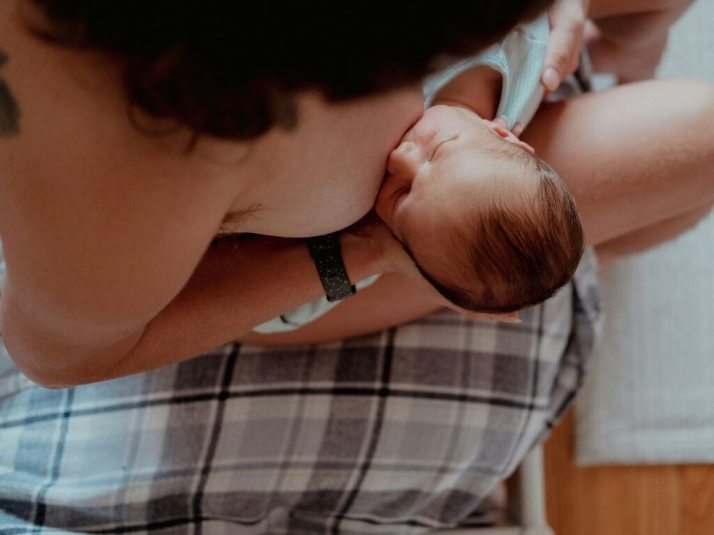 Nő felülnézetből kisbabát szoptat ágy szélén ülve