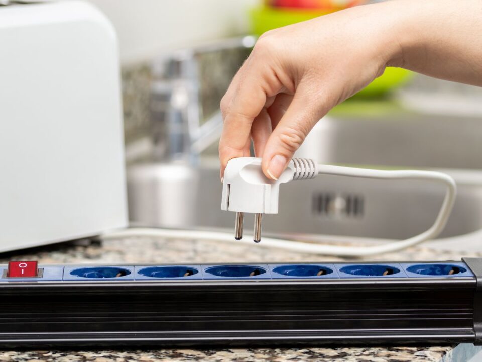 Egy kéz éppen kihúz egy fehér elektromos dugót egy hosszabbítóból a konyhapulton, a háttérben háztartási gépek láthatók.