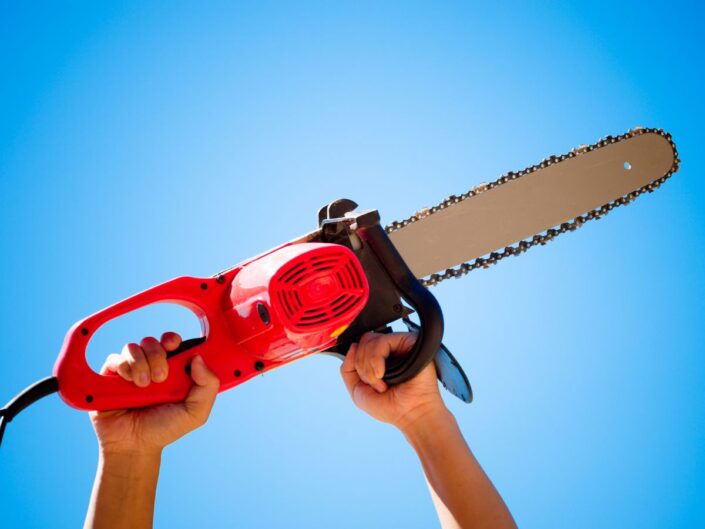 Egy piros, modern láncfűrészt tart két kéz a magasba, tiszta kék ég előtt; a hosszú, fémláncos penge vízszintesen előre nyúlik.