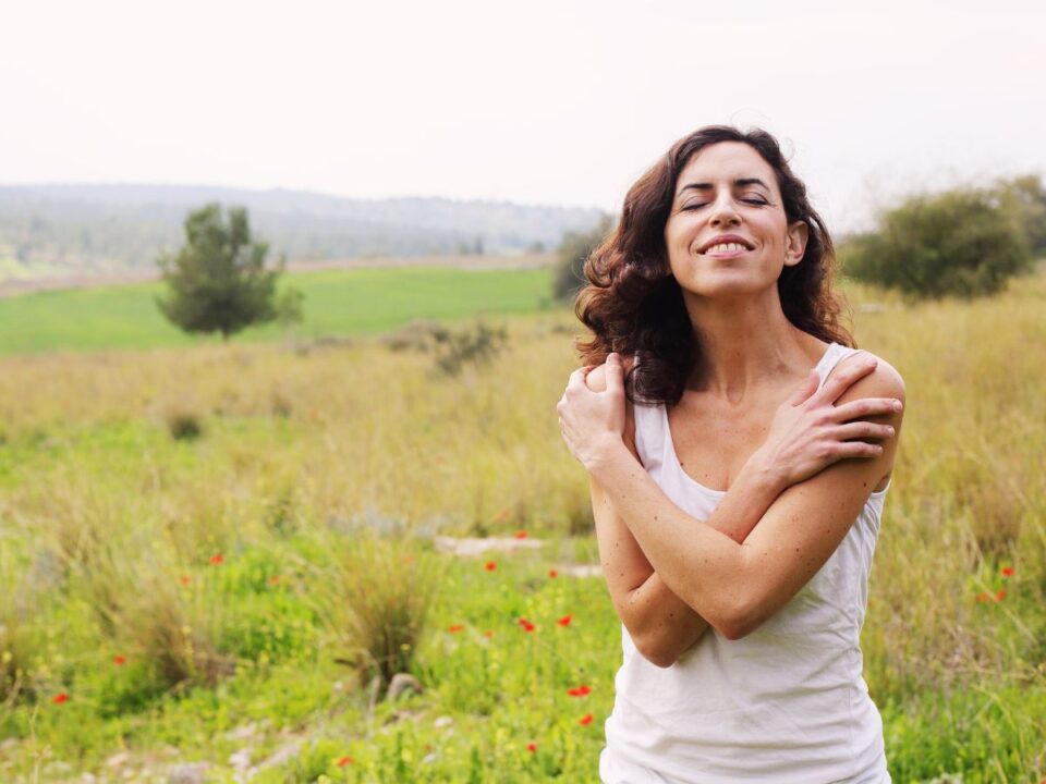 Sötétebb hajú nő egy mezőn áll, lehunyt szemmel, karjait önmagát átölelve tartja. Körülötte zöld táj, távolban dombok, arca békét és megkönnyebbülést tükröz.