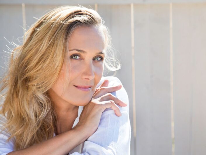 Világos bőrű, szőke nő közelről, természetes fényben. Fehér ruhában ül egy kerítés előtt, enyhén mosolyog, tekintete nyugodt és magabiztos.