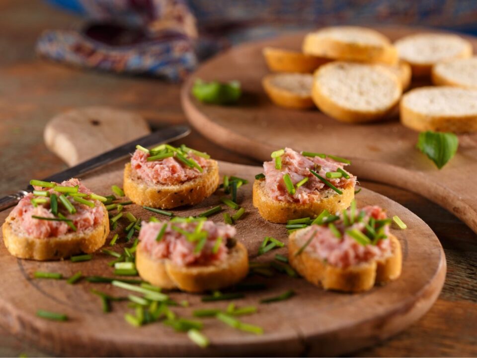 Pirított bagettszeleteken rózsaszínes kenőmájas falatok, apróra vágott snidlinggel megszórva, fa tálon tálalva, barátságos, otthonos hangulatban.