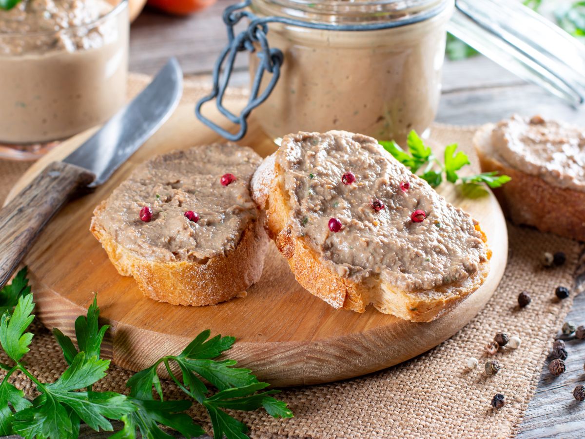 Rusztikus fa deszkán szeletelt kenyér, rajta krémes májpástétom, mellette üvegben még több kenhető májas, petrezselyemmel és borsszemekkel körítve.