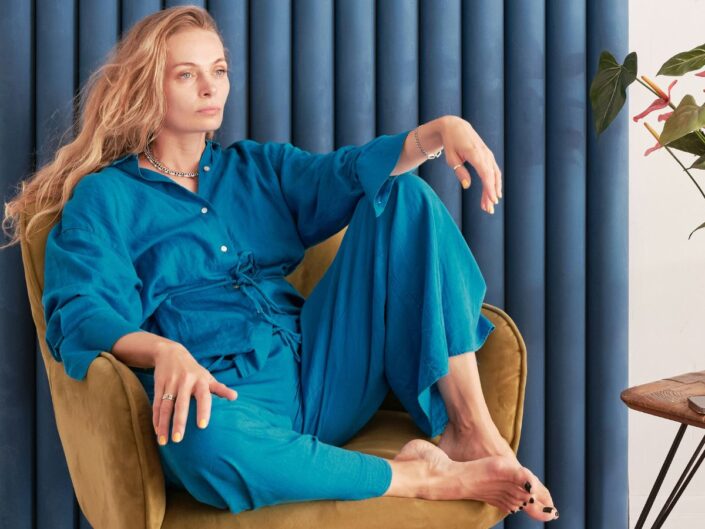 Szép középkorú, hosszú, szőke hajú nő kék overálban felhúzott lábakkal ül sárga fotelben, háta mögött kék függöny