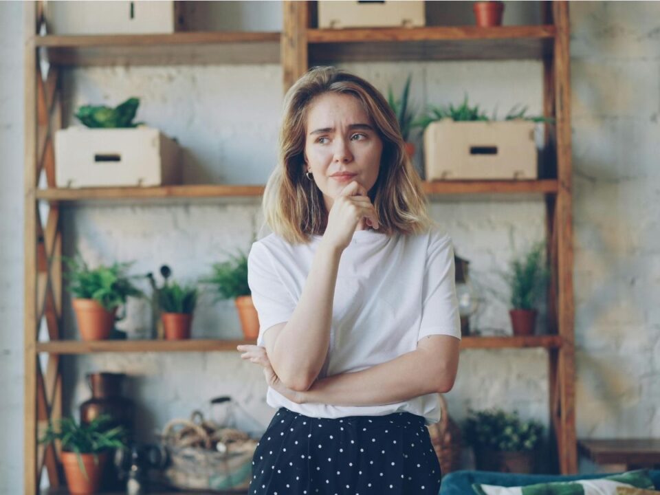 stresszes lány áll a lakásban egy növényekkel dobozokkal teli polc előtt