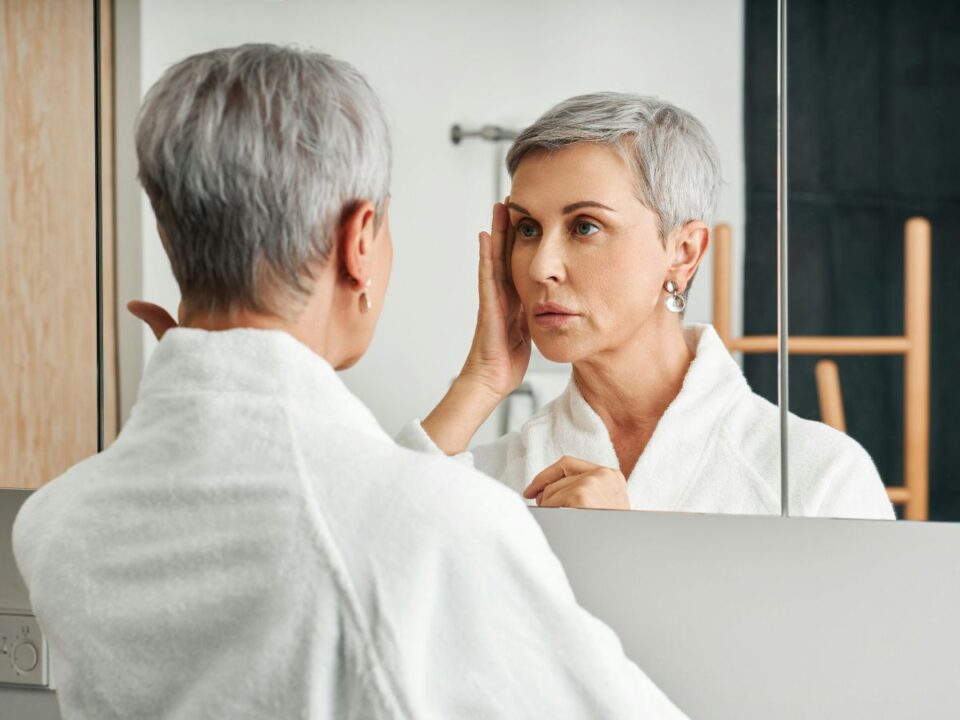 Rövid, ősz hajú nézi magát a tükörben fehér blúzban