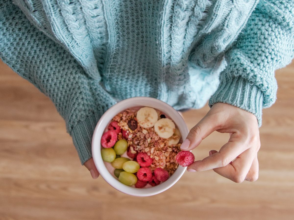 Nő világoskék pulcsiban egy tál rostban gazdag ételt tart a kezében, felülnézetből fotózott kép