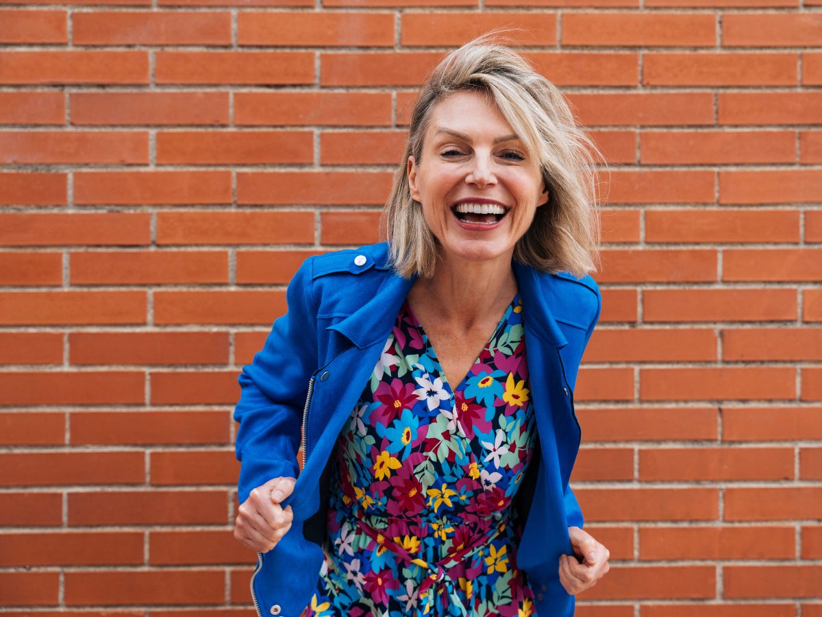 Stílusos 50 feletti, szőke bob hajú nő kék dzsekiben, színes, virágos ruhában nevetve hajol előre téglafal előtt