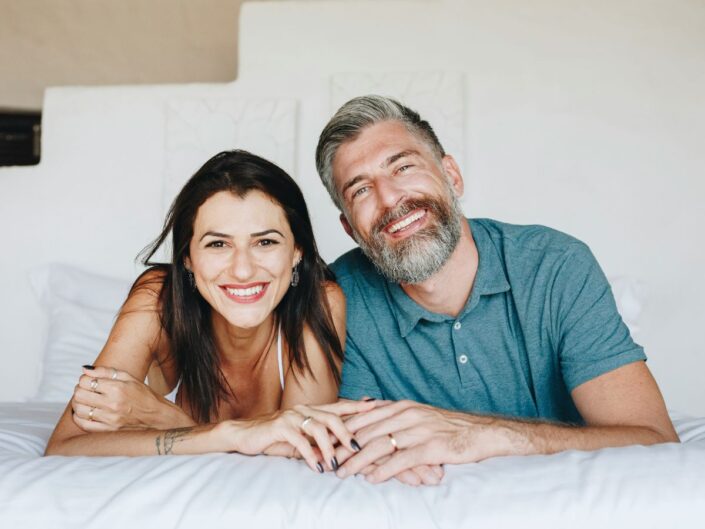 Negyvenes éveiben járó nő és férfi boldogan néz ágyon hasalva a kamerába