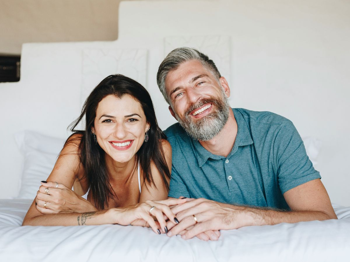 Negyvenes éveiben járó nő és férfi boldogan néz ágyon hasalva a kamerába