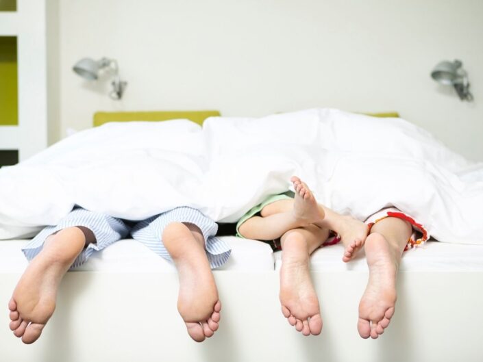 ágyban fekszenek a szülők gyerekkel csak a lábuk lóg ki a takaró alól