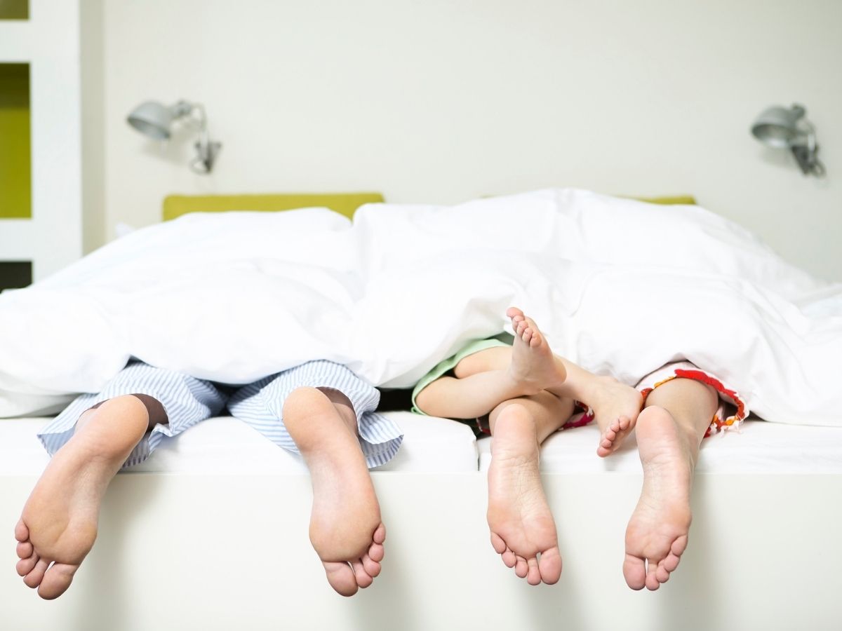 ágyban fekszenek a szülők gyerekkel csak a lábuk lóg ki a takaró alól